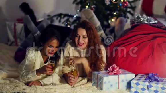 年轻的女朋友一个黑发女人和一个红头发的女孩躺在圣诞树附近的地毯上喝着Shompanskoe视频的预览图