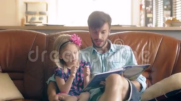 一位年轻的父亲和他快乐的金发小女儿正在餐厅的皮革沙发上读一个故事4K视频的预览图