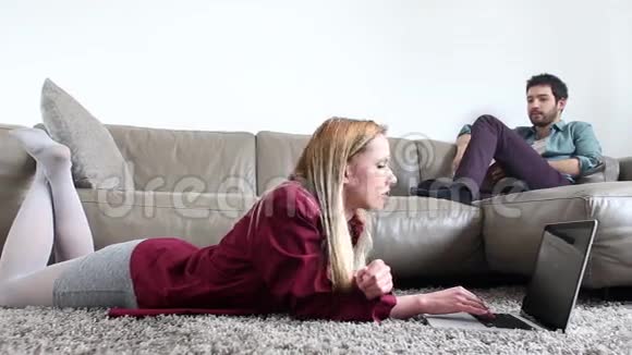 女人躺在地毯上看笔记本电脑而男人坐在沙发上看书视频的预览图