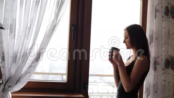 迷人的年轻女士休息在家的窗户前喝一杯茶或咖啡视频的预览图