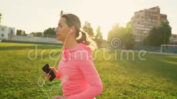 日落时分戴着耳机和智能手机的女人穿过体育场视频的预览图
