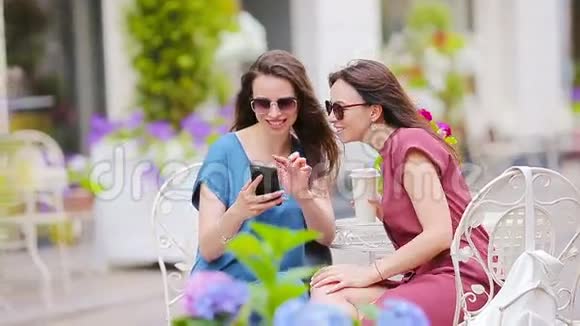 两个年轻女孩在户外咖啡馆用智能手机自拍两个女人在买完东西后拿着袋子坐着视频的预览图
