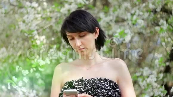 公园里有智能手机的年轻女人春天在一棵开花的树的背景下视频的预览图