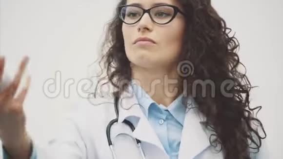 年轻漂亮的女医生穿着医用礼服在这段时间里她的背景是灰色的手里拿着一个白色的视频的预览图