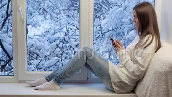 坐在窗台上用智能手机的漂亮女孩冬天在外面视频的预览图