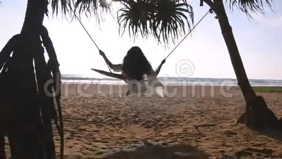 年轻快乐的女人穿着泳衣和衬衫在热带海洋海滩的秋千上放松坐在秋千上的漂亮女孩视频的预览图