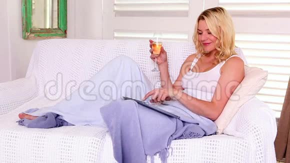 一个漂亮的金发女郎带着平板电脑和葡萄酒在沙发上放松视频的预览图
