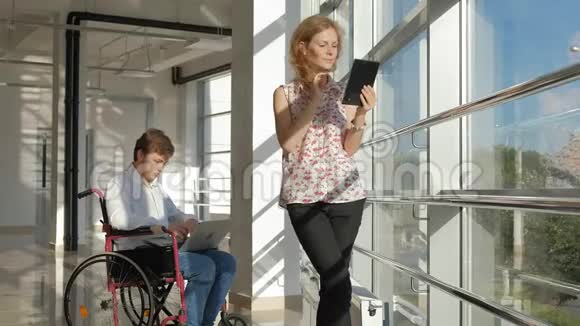 一位坐轮椅的残疾商人在窗户旁手提电脑旁边的女士使用平板电脑视频的预览图