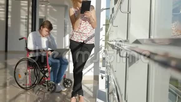 一位坐轮椅的残疾商人在窗户旁手提电脑旁边的女士使用平板电脑视频的预览图