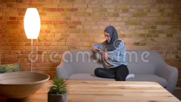 在舒适的家中坐在沙发上用手机拍摄穆斯林年轻女性戴头巾的特写镜头视频的预览图