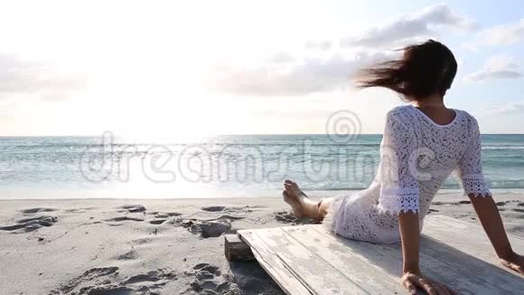 坐在海边的年轻女子在风中看着黎明时的地平线身穿白色蕾丝连衣裙和白色视频的预览图