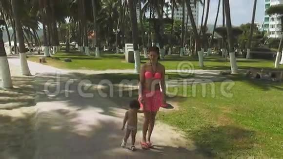 年轻漂亮的女人穿着粉红色的泳衣带着一个婴儿在公园和瓦维的背景下漫步在水边视频的预览图