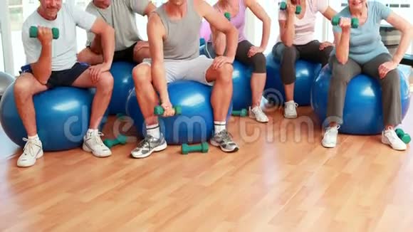 健身小组坐在健身球上举起手中的重物对着镜头微笑视频的预览图