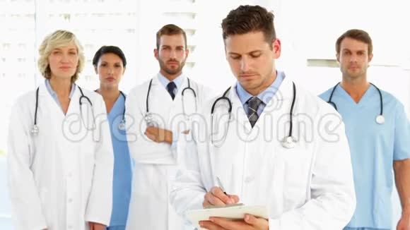 当工作人员站在他身后时严肃的医生在剪贴板上写字视频的预览图