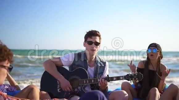 年轻人在一群朋友中弹吉他他们坐在沙滩上的安乐椅上在一个夏天的晚上唱歌视频的预览图