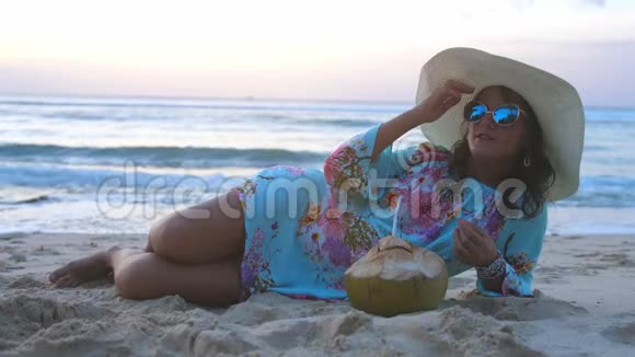 年轻的旅游美女戴着墨镜和帽子躺在热带天堂沙滩上喝椰子汁视频的预览图