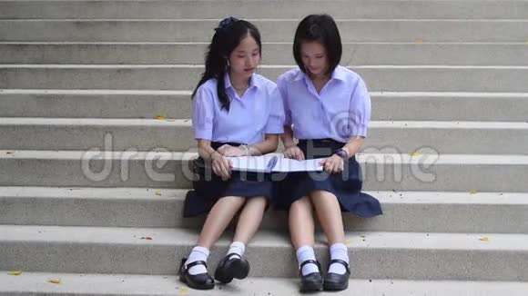 可爱的亚洲泰国高中女生夫妇穿着校服坐在楼梯上讨论和阅读家庭作业或考试视频的预览图