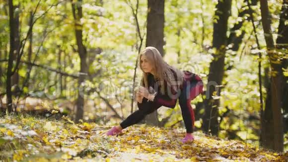 女人在户外做健身运动在秋林中伸展的女性户外健身的苗条女孩蹲视频的预览图