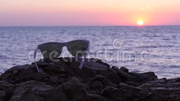 太阳眼镜躺在大海日落背景的石头上视频的预览图