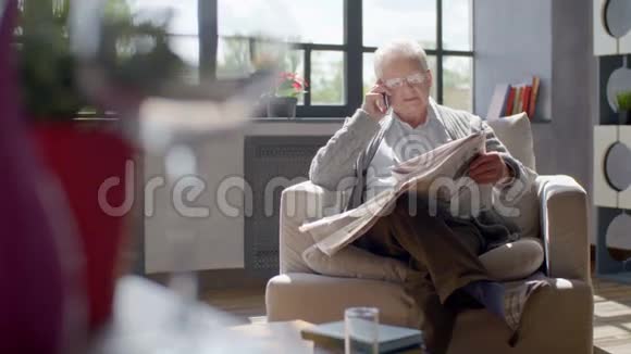 老人坐在椅子上在现代化的公寓里看报纸和打电话视频的预览图
