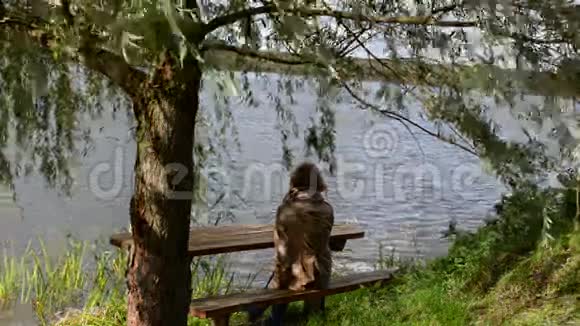 女子坐木凳柳树动风湖放松视频的预览图