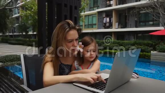 年轻的母亲和小女儿在游泳池附近用笔记本电脑慢动作聊天视频的预览图