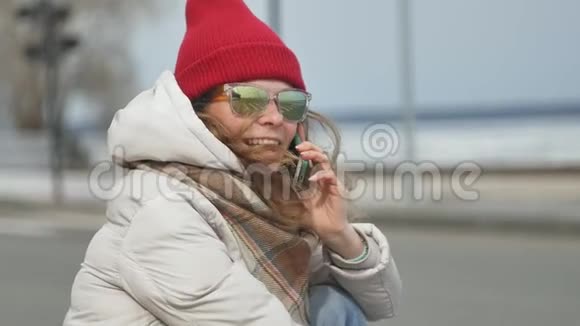 年轻漂亮的女人戴着一顶红色帽子穿着运动温暖的衣服和滚筒坐在柏油路上谈论着视频的预览图