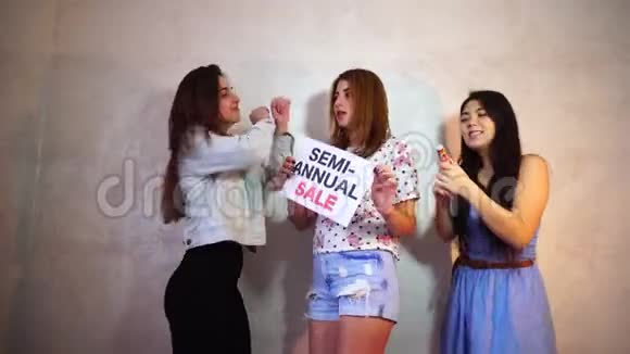 三个时尚的女性朋友摆姿势带标志并呼吁购物站在房间的背景下轻墙视频的预览图