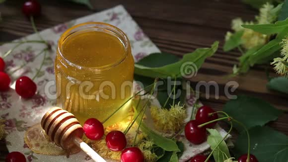 打开玻璃罐的液体蜂蜜和蜂蜜迪珀一束林登花和红樱桃在木制表面阳光的光芒视频的预览图