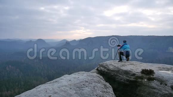 摄影师穿着蓝色夹克穿着温暖的衣服站在岩石的视点上拍摄朦胧的早晨景观在岩石里远足视频的预览图