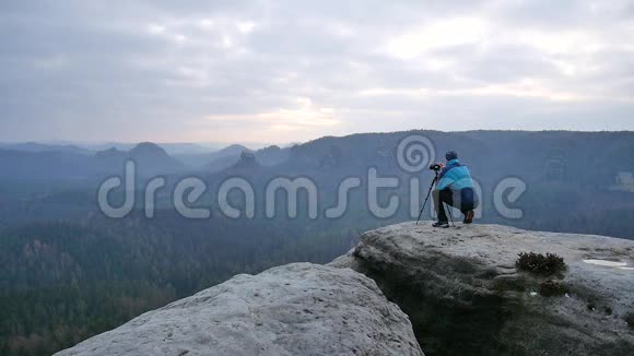 穿着蓝色夹克衫的摄影师站在岩石的视点上拍摄朦胧的早晨景观在岩石里远足视频的预览图