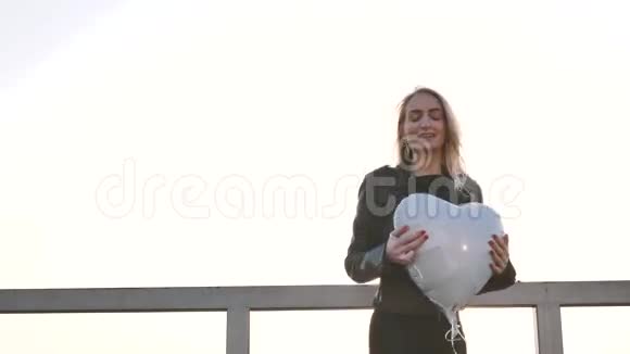 夕阳西下一个年轻漂亮的女孩带着一个心形的气球站在屋顶的栏杆附近视频的预览图