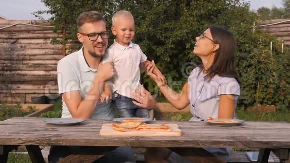 在花园里野餐的年轻幸福家庭妈妈爸爸和儿子吃披萨店视频的预览图
