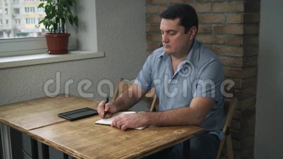 成年男性坐在一家有日记和平板电脑的咖啡馆里在笔记本上记笔记视频的预览图
