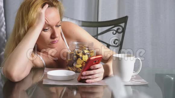 饥肠辘辘的伤心女人晚上坐在客厅的桌子旁吃着肝脏用着智能手机视频的预览图