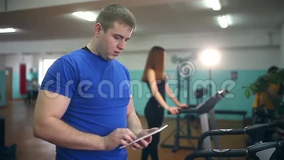 男教练在健身房女孩带着平板电脑在跑步机上健身健康的生活方式运动视频的预览图