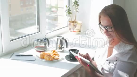 年轻漂亮的女孩在阳光明媚的日子里用平板电脑吃早餐视频的预览图