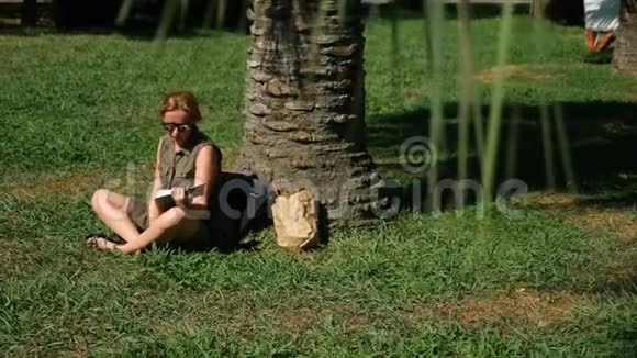 金发女人在公园里看书坐在草地上树下4k慢镜头视频的预览图
