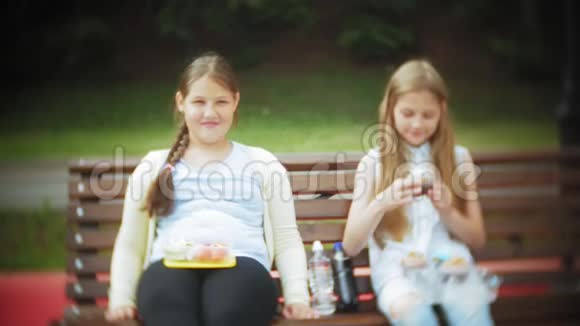 两个小女孩苗条又胖吃健康有害的食物蔬菜和蛋糕坐在咖啡馆的长凳上视频的预览图
