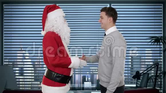 圣诞老人和年轻的商人在现代化的办公室里握手看着摄像机股票录像视频的预览图
