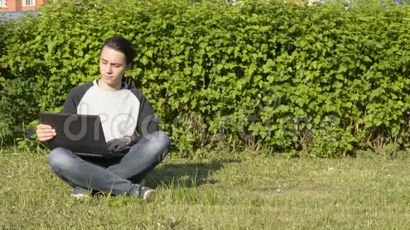 英俊的年轻人坐在公园的草地上用笔记本电脑工作视频的预览图