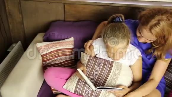 年轻的妈妈在睡前在沙发上的客厅里大声地给女儿读一本书视频的预览图