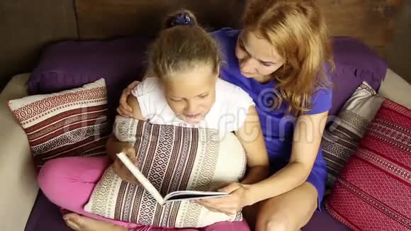 年轻的妈妈在睡前在沙发上的客厅里大声地给女儿读一本书视频的预览图