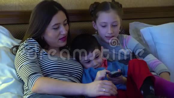 一位年轻的母亲带着两个孩子躺在旅馆的床上看电视使用电视遥控器视频的预览图