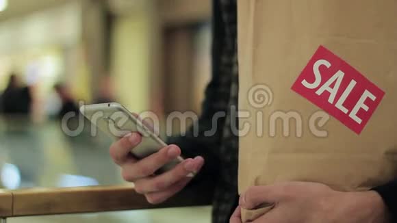 男士一只手拿着带有销售标签的棕色纸袋在商场里使用智能手机视频的预览图