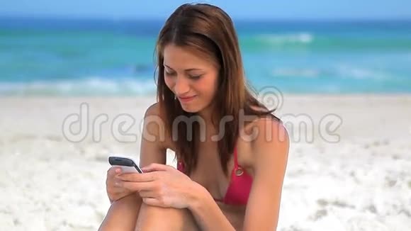 微笑的黑发女人坐在沙滩上发短信视频的预览图