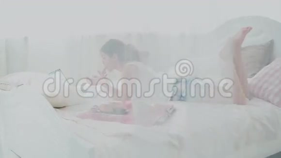 年轻女子使用无线技术躺在家里的白色床上女孩在早餐时在笔记本电脑上聊天视频的预览图
