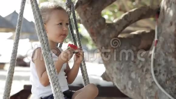 小可爱的小女孩坐在秋千上在沙滩上吃红色的西瓜视频的预览图