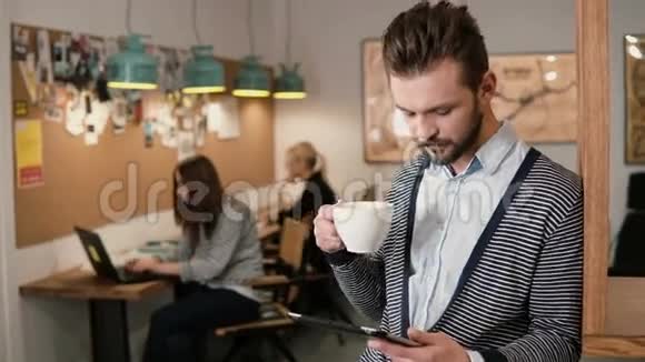 年轻英俊的胡须男在现代的创业办公室里使用触摸屏平板电脑和喝咖啡视频的预览图