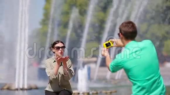 男朋友坐在喷泉的背景下给他的朋友拍照年轻人在女人的照片上视频的预览图